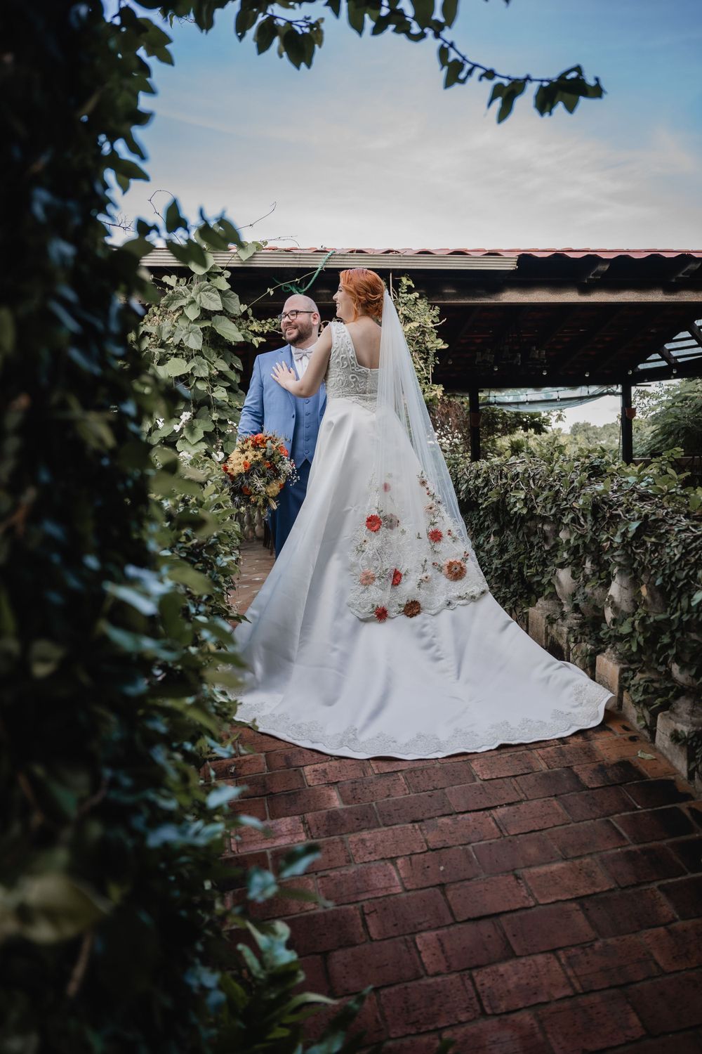 A couple in wedding attire stands on brick pavers surrounded by greenery, with the bride wearing a floral-adorned white gown.
