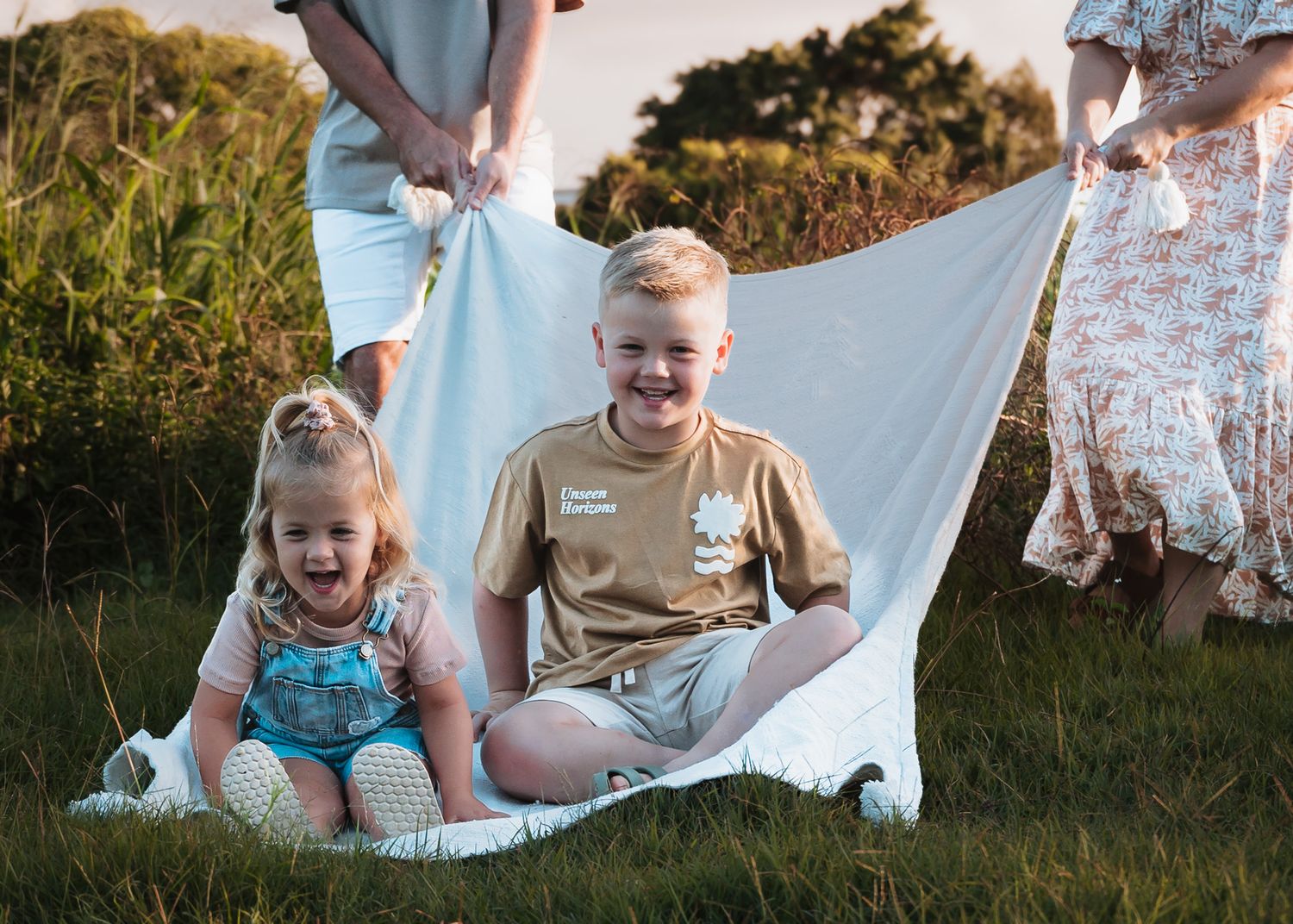Two children sit together on white blanket during outdoor photo session.