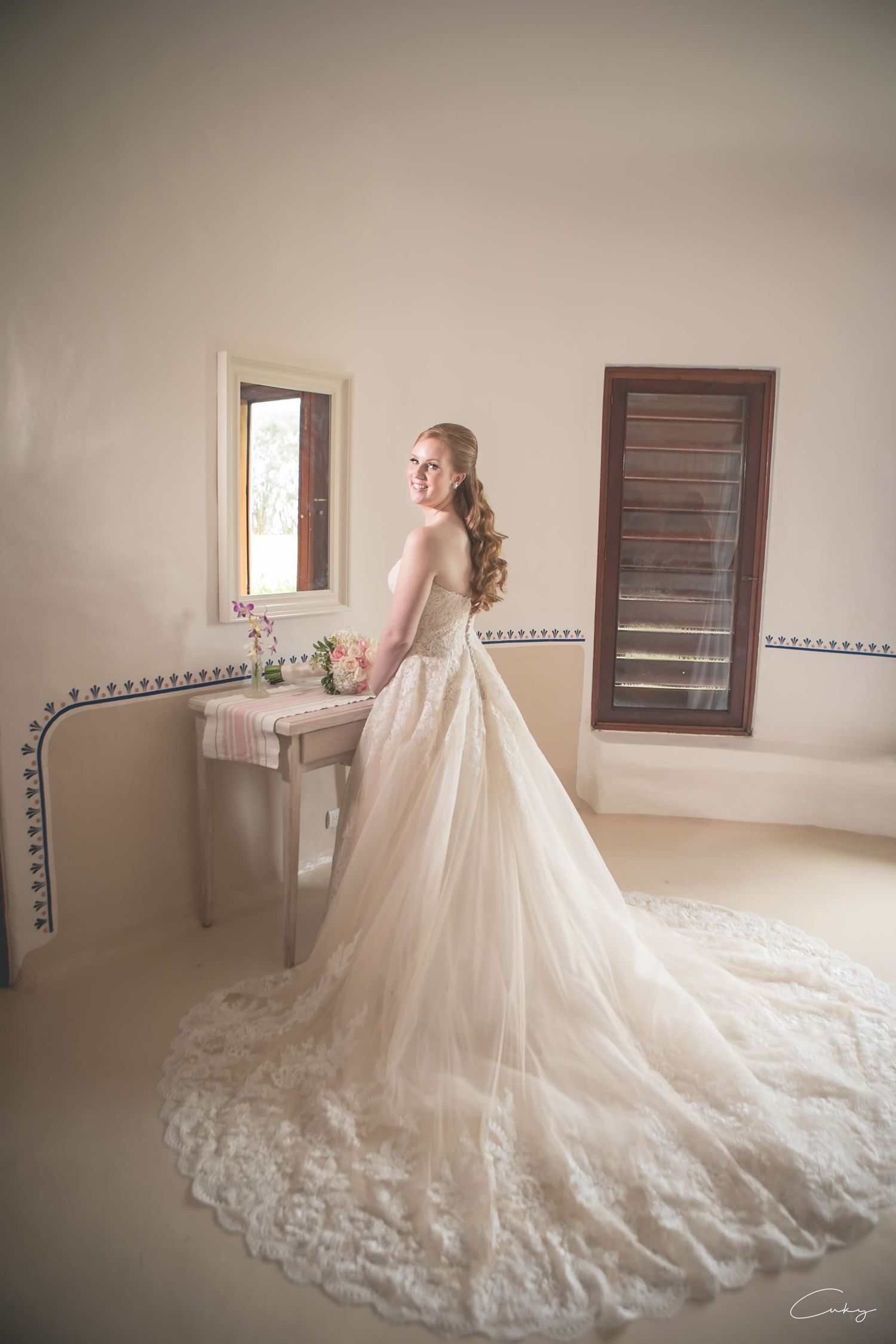 Elegant bride in lace wedding gown with long train poses by window in getting ready room.