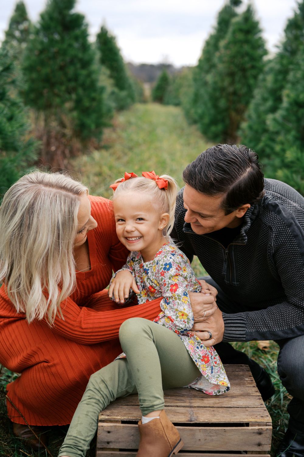 Candid family moment on wooden crate surrounded by evergreen trees.