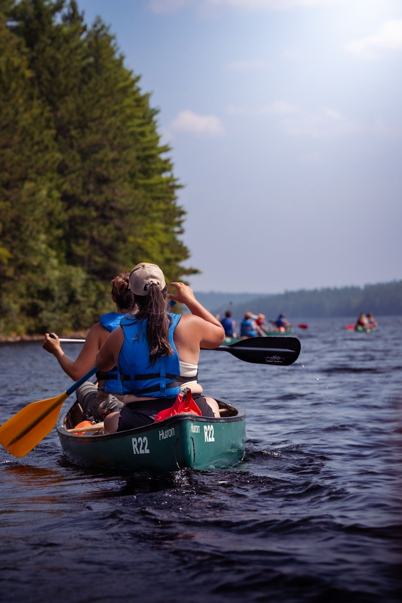 Kayakers paddle across a serene lake surrounded by pine trees on a summer afternoon while wearing blue life vests.