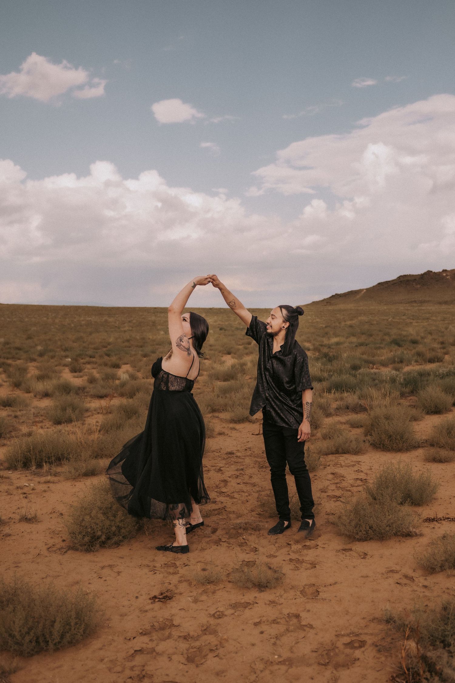 A couple dances together in a desert landscape with cloudy skies. engagement photographer albuquerque. New Mexico wedding photographer LGBT.