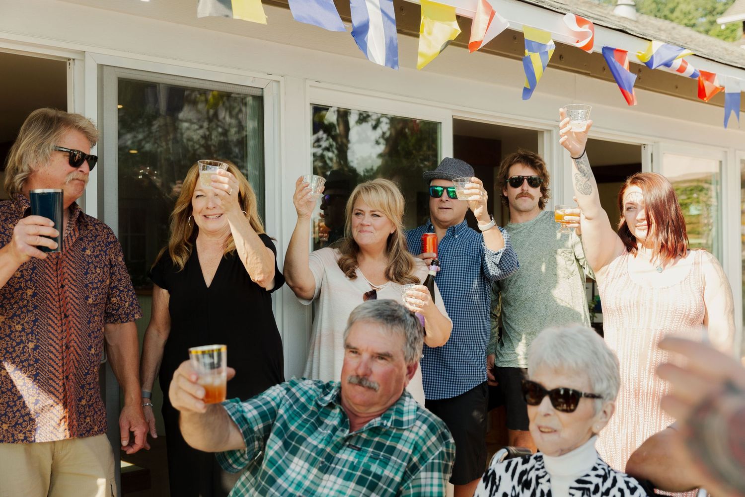 A group of party guests raise their glasses in a celebratory toast on an outdoor porch decorated with colorful flags.