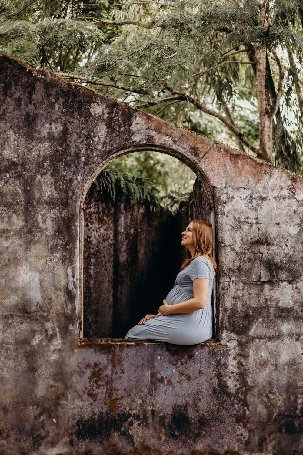 A person in a gray dress sits peacefully in a stone archway framed by overhanging trees.