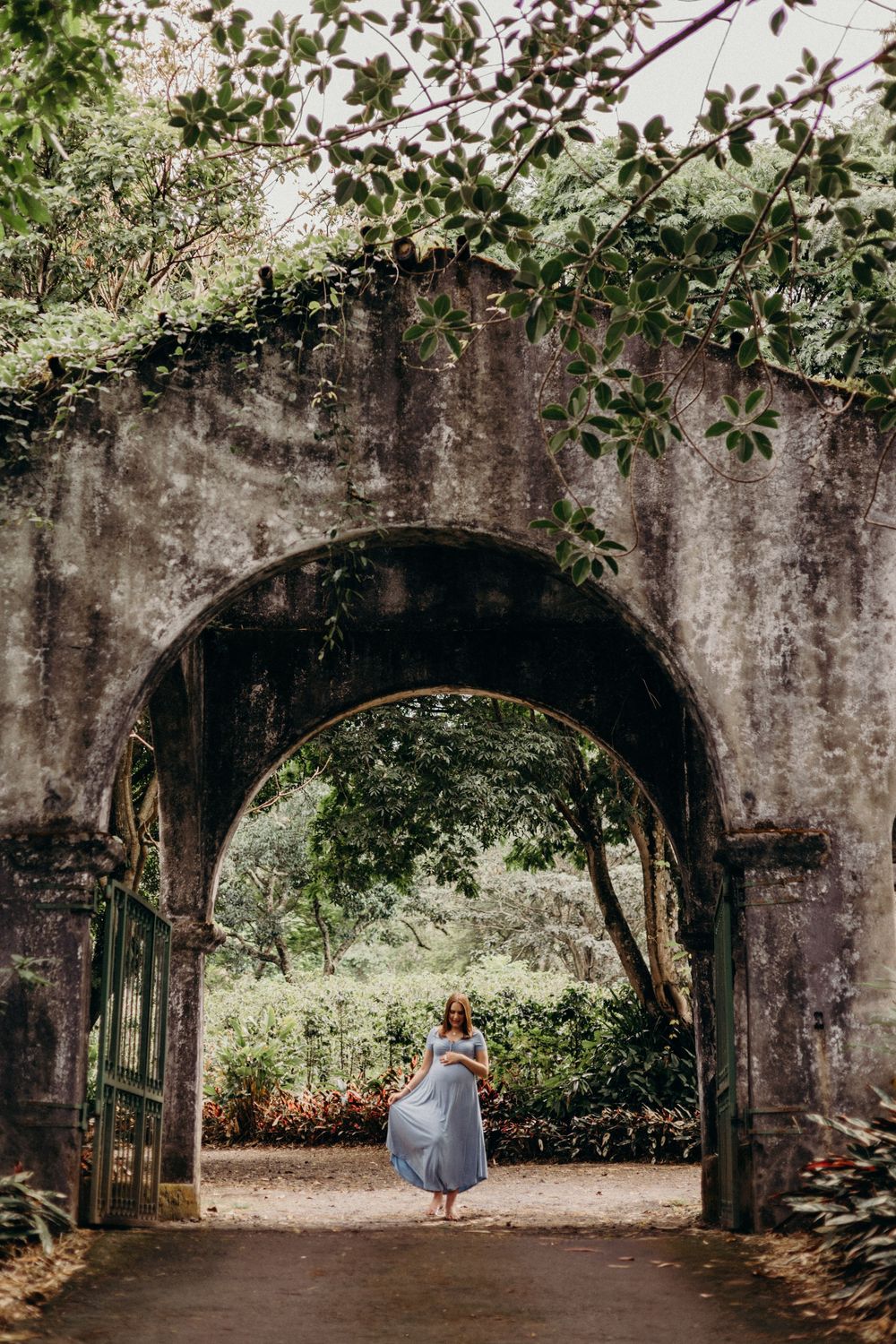 A stone archway covered in lush green foliage frames a path where someone stands in a flowing blue dress.