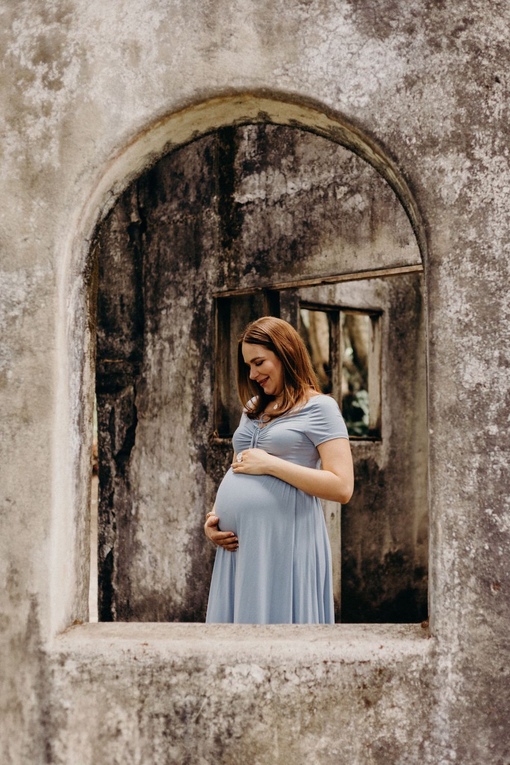 A pregnant woman in a light blue dress stands in an arched stone window frame of an old weathered building.