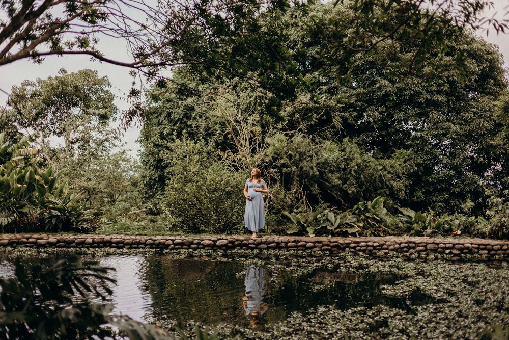 A figure stands on a wooden footbridge over a tranquil pond surrounded by lush greenery and trees.