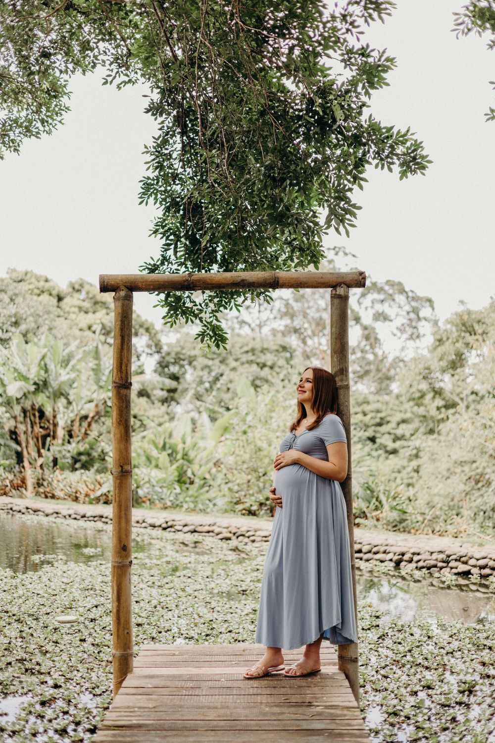 A serene maternity photo showing an expectant mother in a grey dress standing under a wooden arch by a pond.