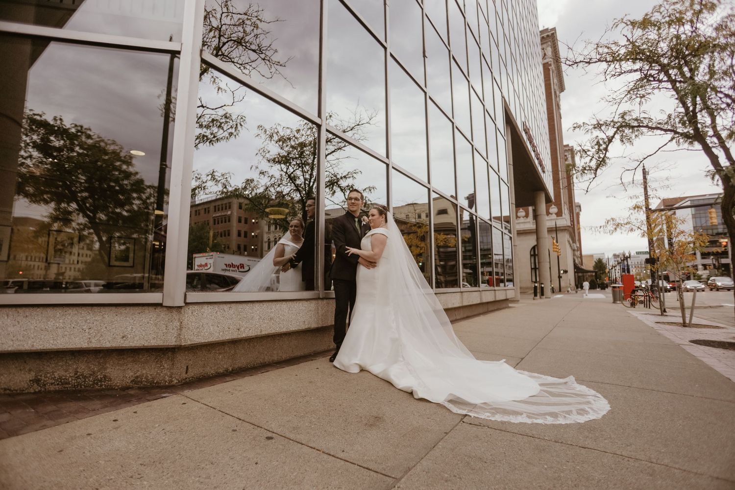 Newlywed couple posing in front of a modern glass building with a flowing white wedding dress train.