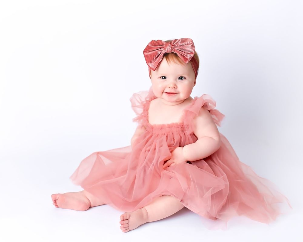 Baby wearing coral pink tulle dress and matching bow headband sits against white backdrop in studio portrait.