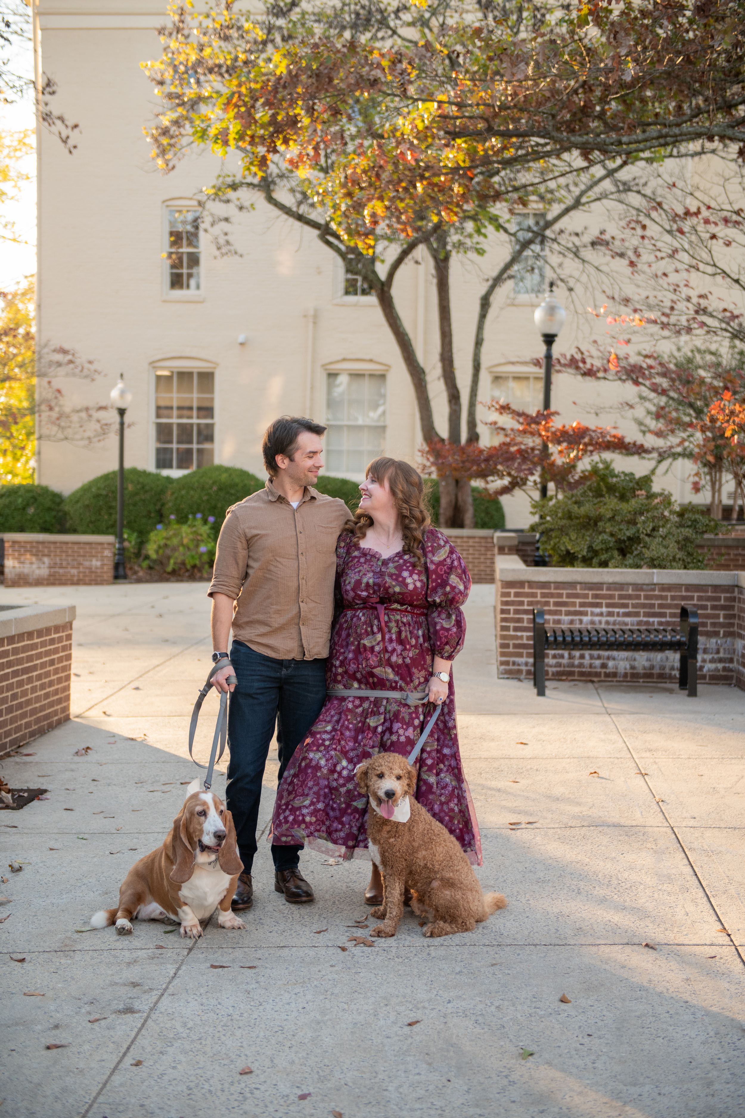A couple sits on a bench with their two dogs during an autumn afternoon on a brick courtyard.