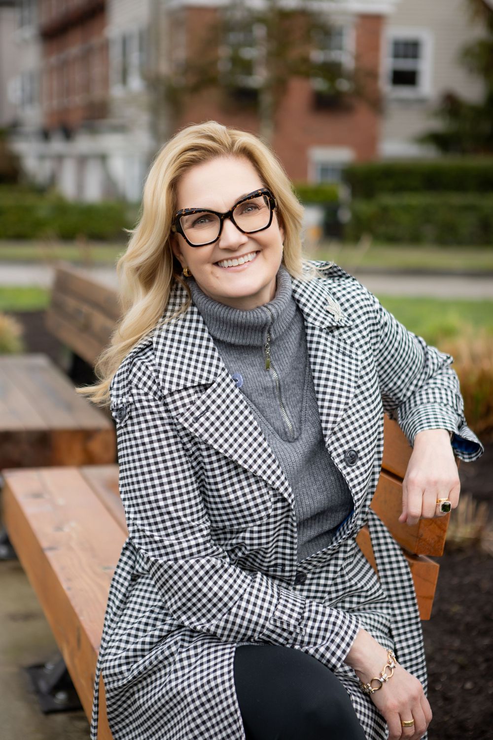 A person wearing glasses and a gray turtleneck sweater with a checkered coat sits smiling on a wooden bench outside.