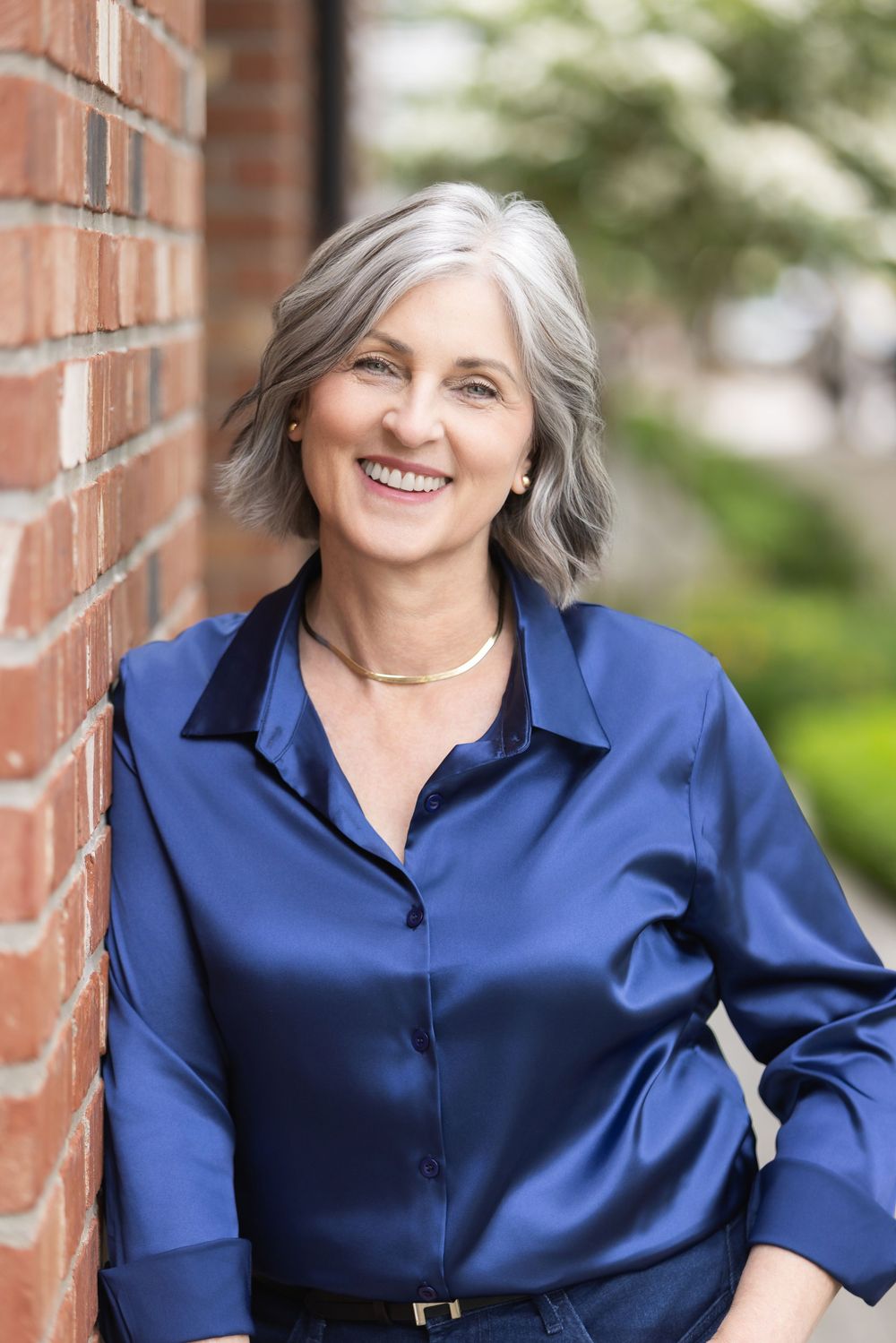 A person in a royal blue button-down shirt poses against a brick wall backdrop with greenery visible in the background.