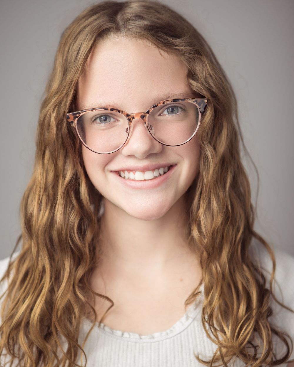 A series of headshots showing someone with wavy brown hair and glasses in various expressions and poses against a gray background.
