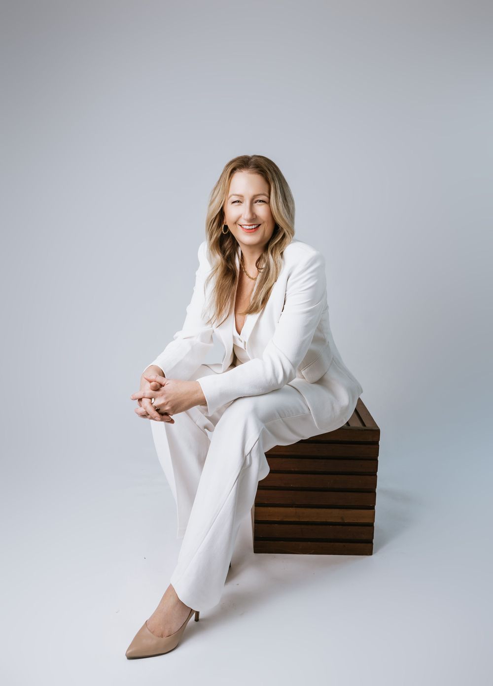 A person wearing a white blazer and pants sits casually on wooden blocks against a light gray background.