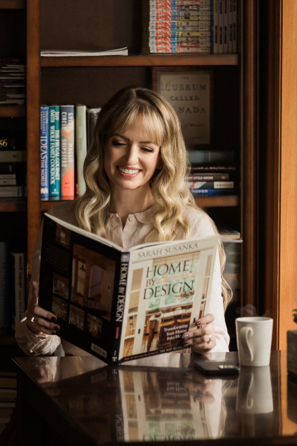 A person in a pink sweater reads home design magazines at a wooden table in a library setting with natural lighting.