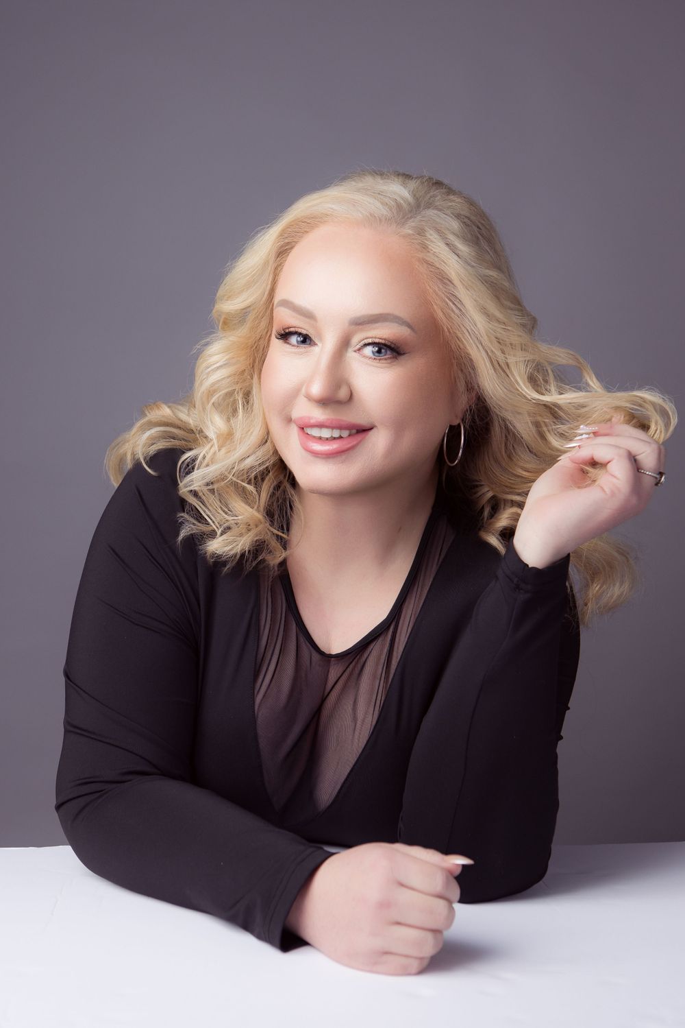 Professional portrait of someone with blonde wavy hair wearing a black top against a gray background, lying on white surface.
