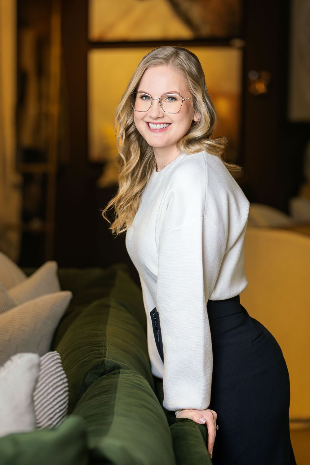 Smiling blonde woman in white blouse and black skirt wearing glasses poses beside a green sofa in elegant interior.