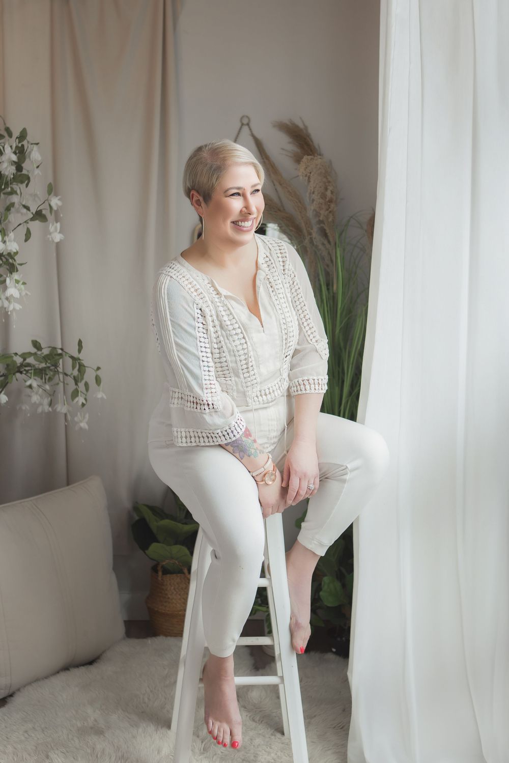 Minimalist indoor portrait with a white lace top, white pants, and soft natural lighting by a window with pampas grass decor.