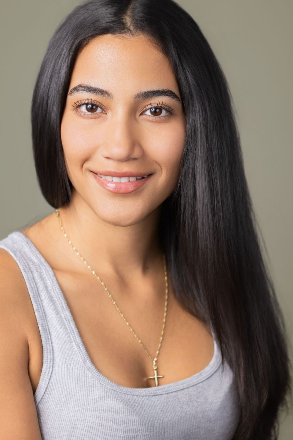 A smiling young woman with long black hair wearing a gray tank top and delicate gold necklace against a neutral background.