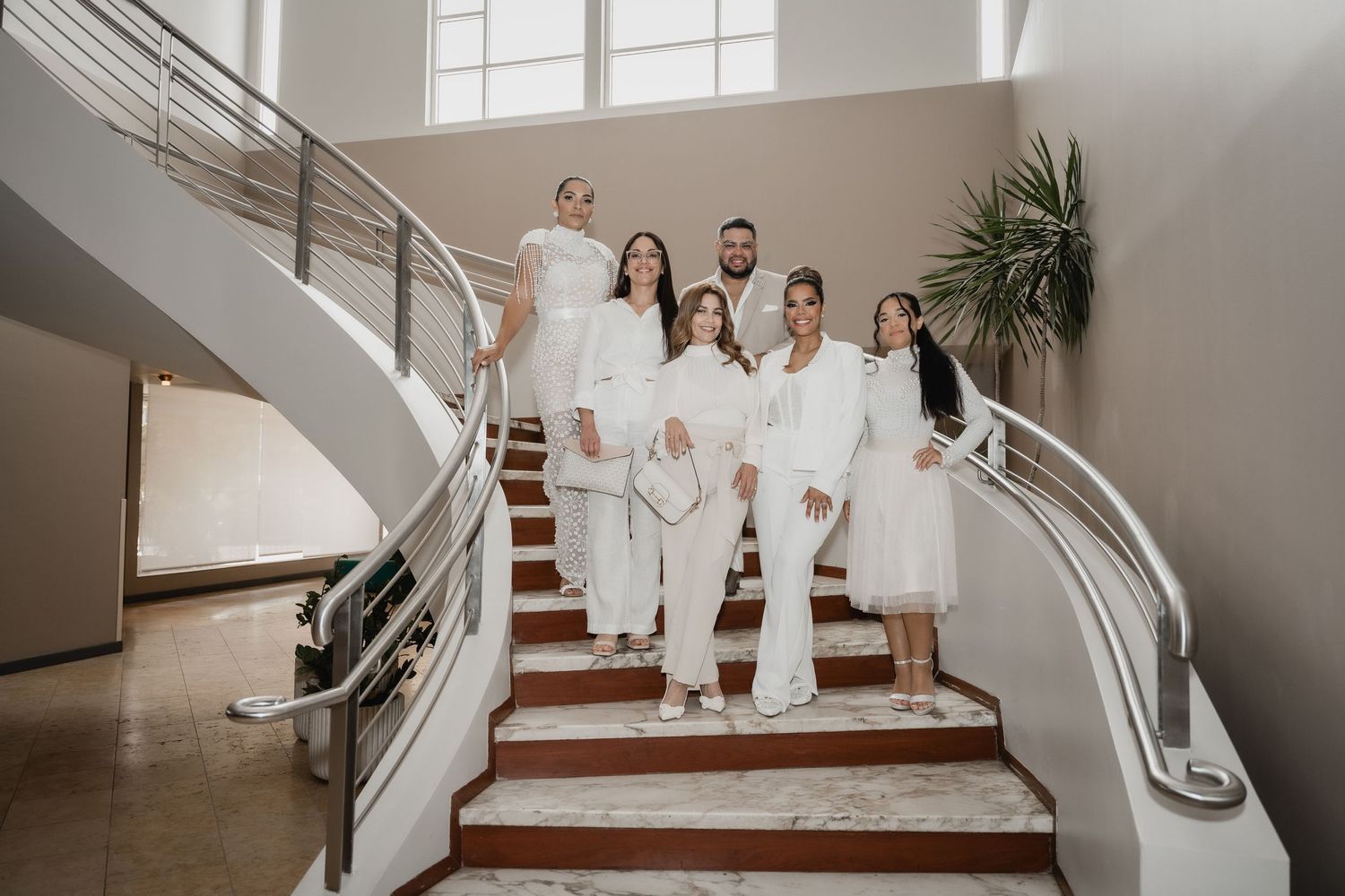 Group in all-white outfits pose on elegant curved staircase with modern interior decor and natural lighting.