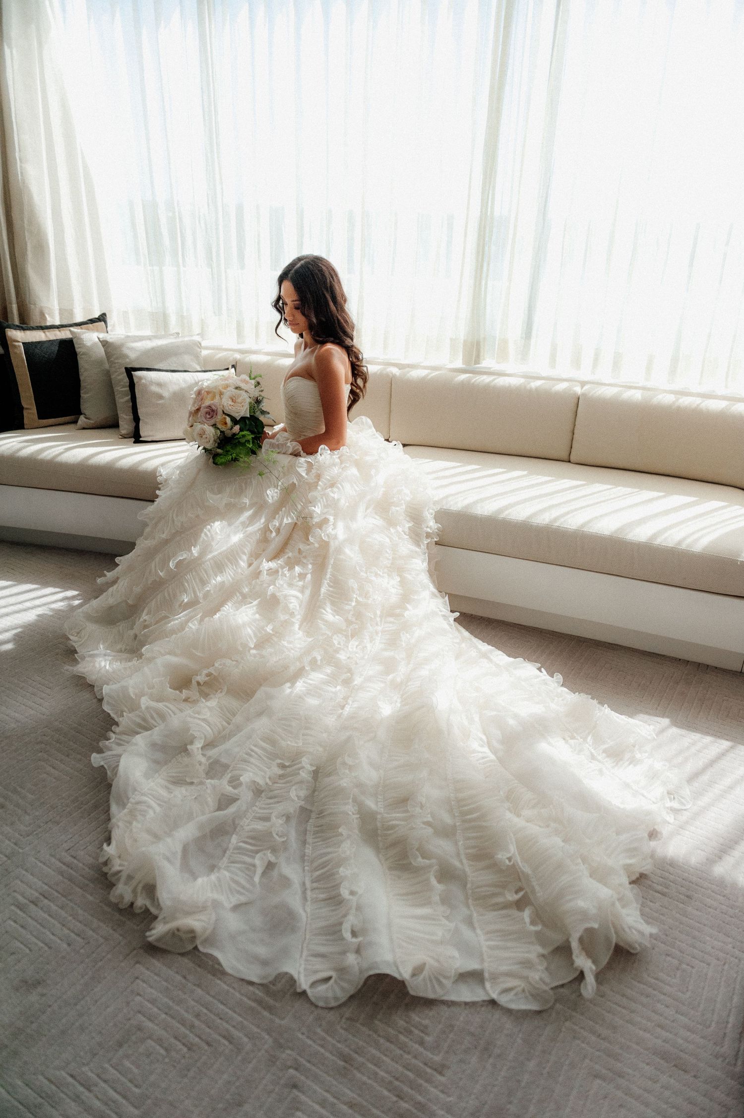 A bride in a stunning layered tulle ball gown with dramatic train poses near sunlit windows in an elegant room.