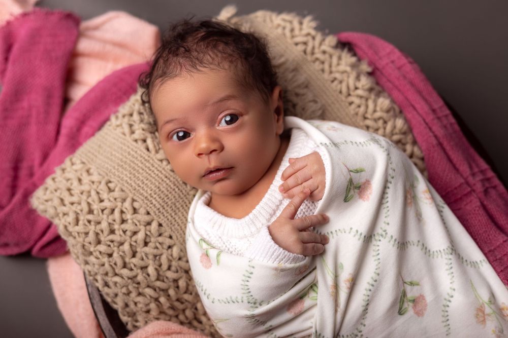 Newborn baby wrapped in white floral swaddle blanket, nestled among pink and beige knit blankets.