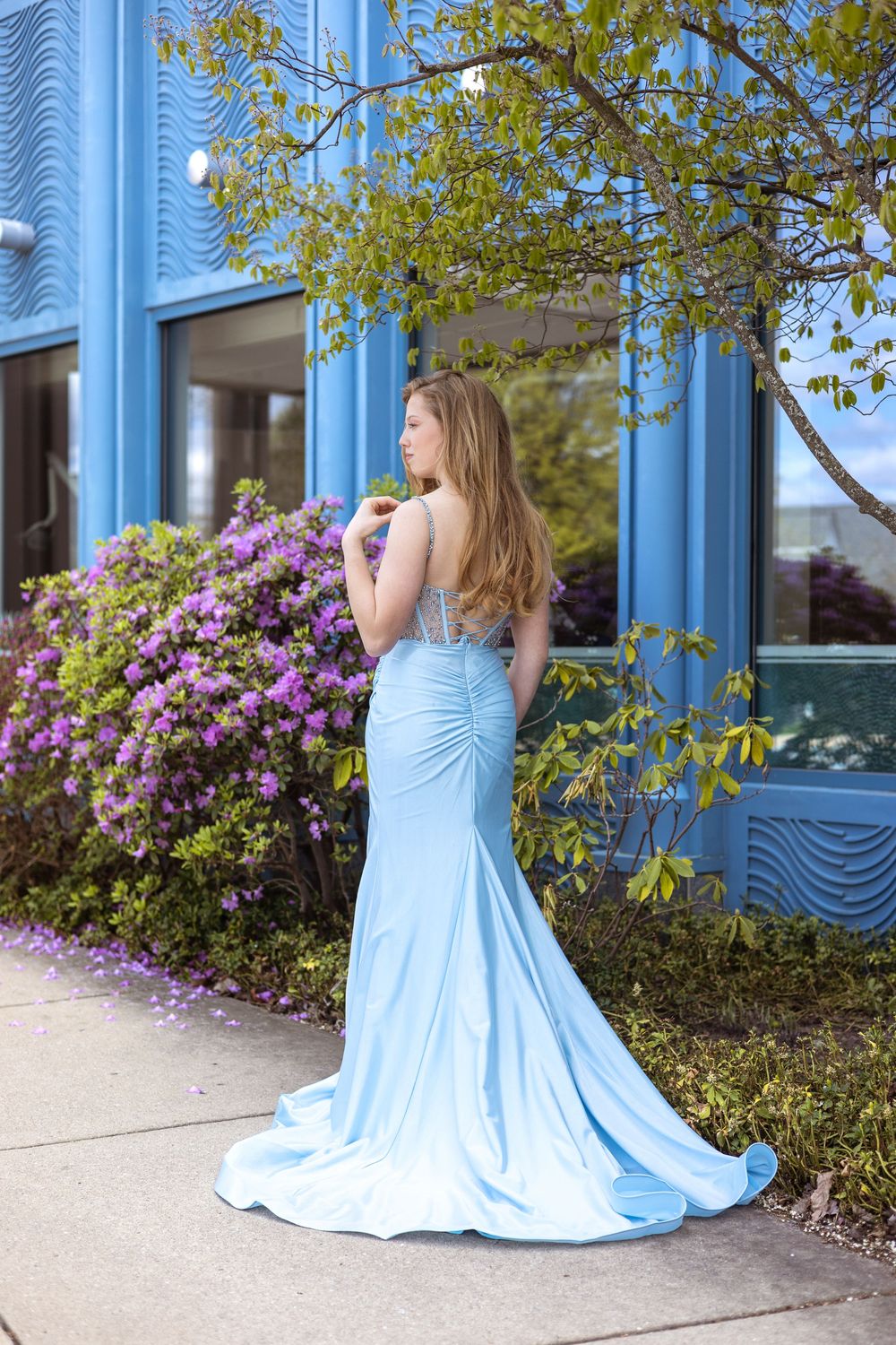 A person in a flowing light blue gown poses in front of a blue building with blooming purple flowers.