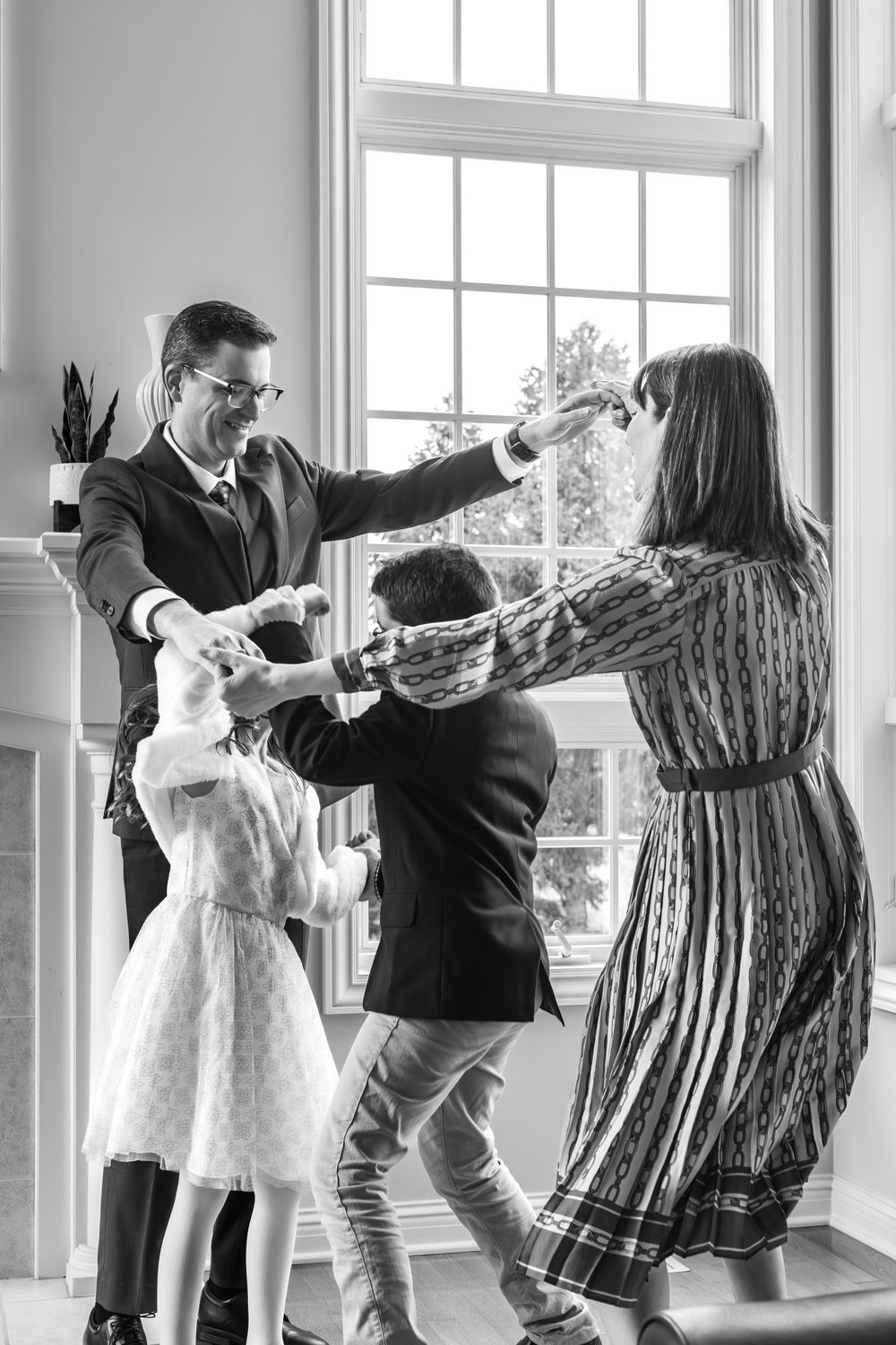 A joyful black and white photo of family members dancing together in a sunlit room with large windows.
