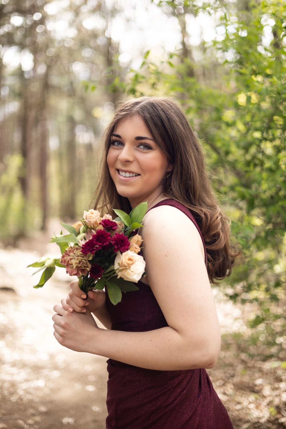 Someone in a burgundy dress holds a colorful bouquet featuring burgundy and peach flowers in a sunlit wooded setting.
