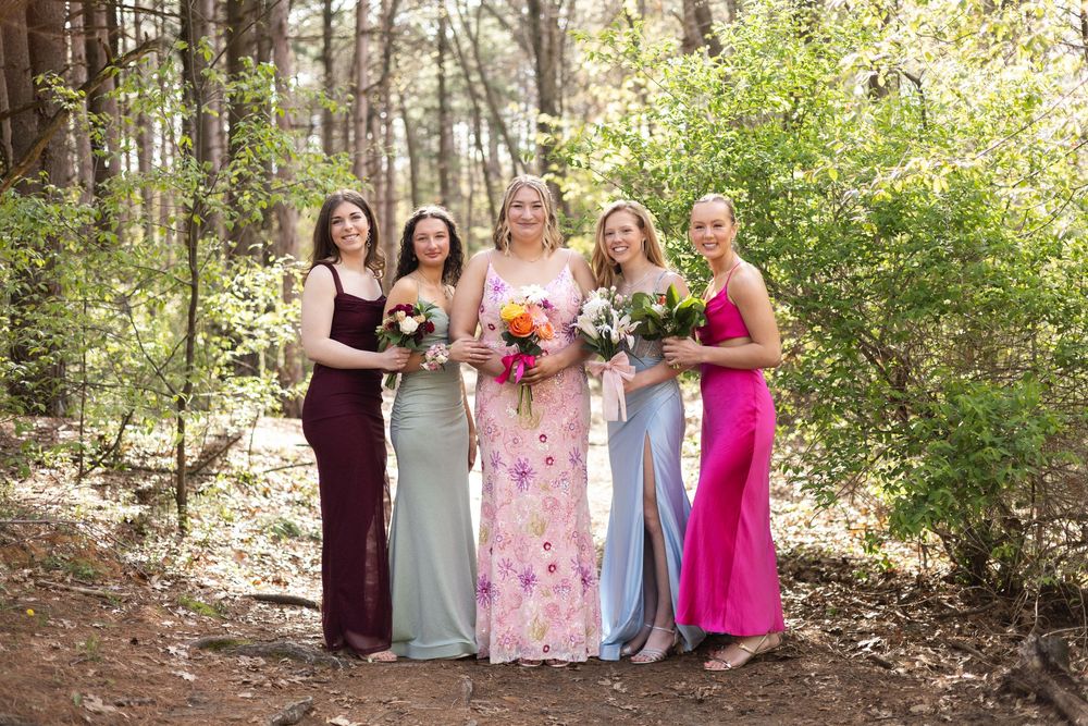 A bridal party poses together in a wooded setting wearing colorful floor-length gowns and holding floral bouquets.