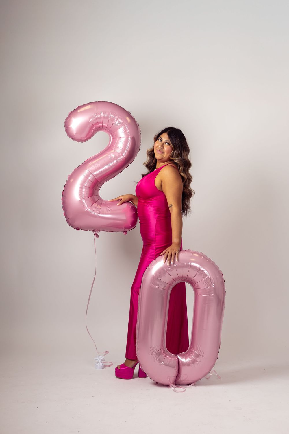Celebratory photo setup with pink metallic number balloons 20 and a figure in a fuchsia dress against a light background.
