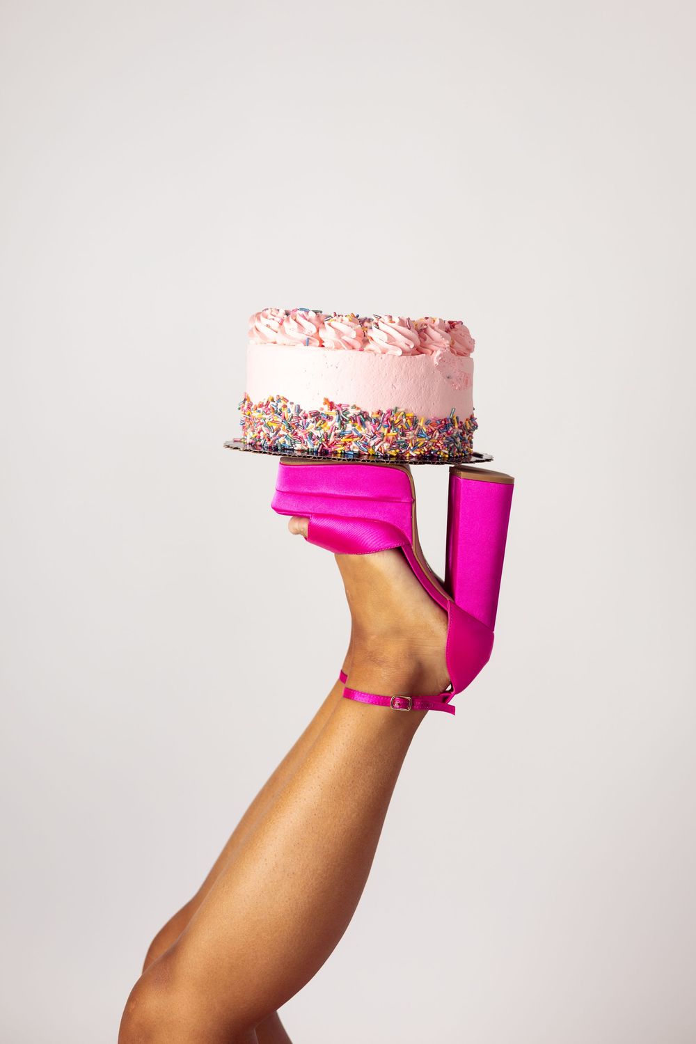 A pink frosted cake with sprinkles balanced on a hot pink high heel shoe against a white background.