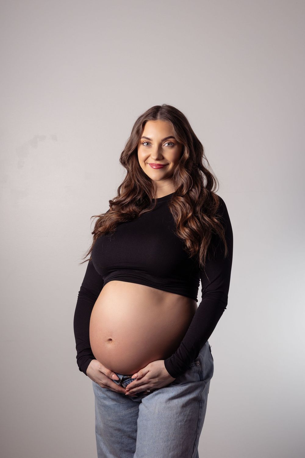 Smiling pregnant woman in black crop top and gray jeans cradling her bare baby bump against a light gray background.