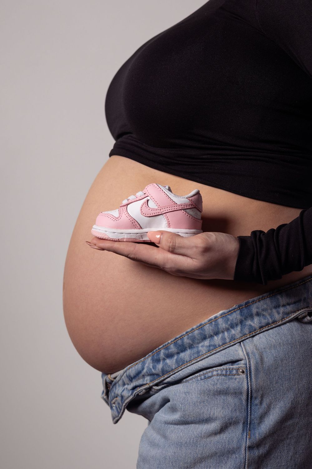 Pregnant woman in black top and jeans holding tiny pink Nike sneakers against her baby bump.