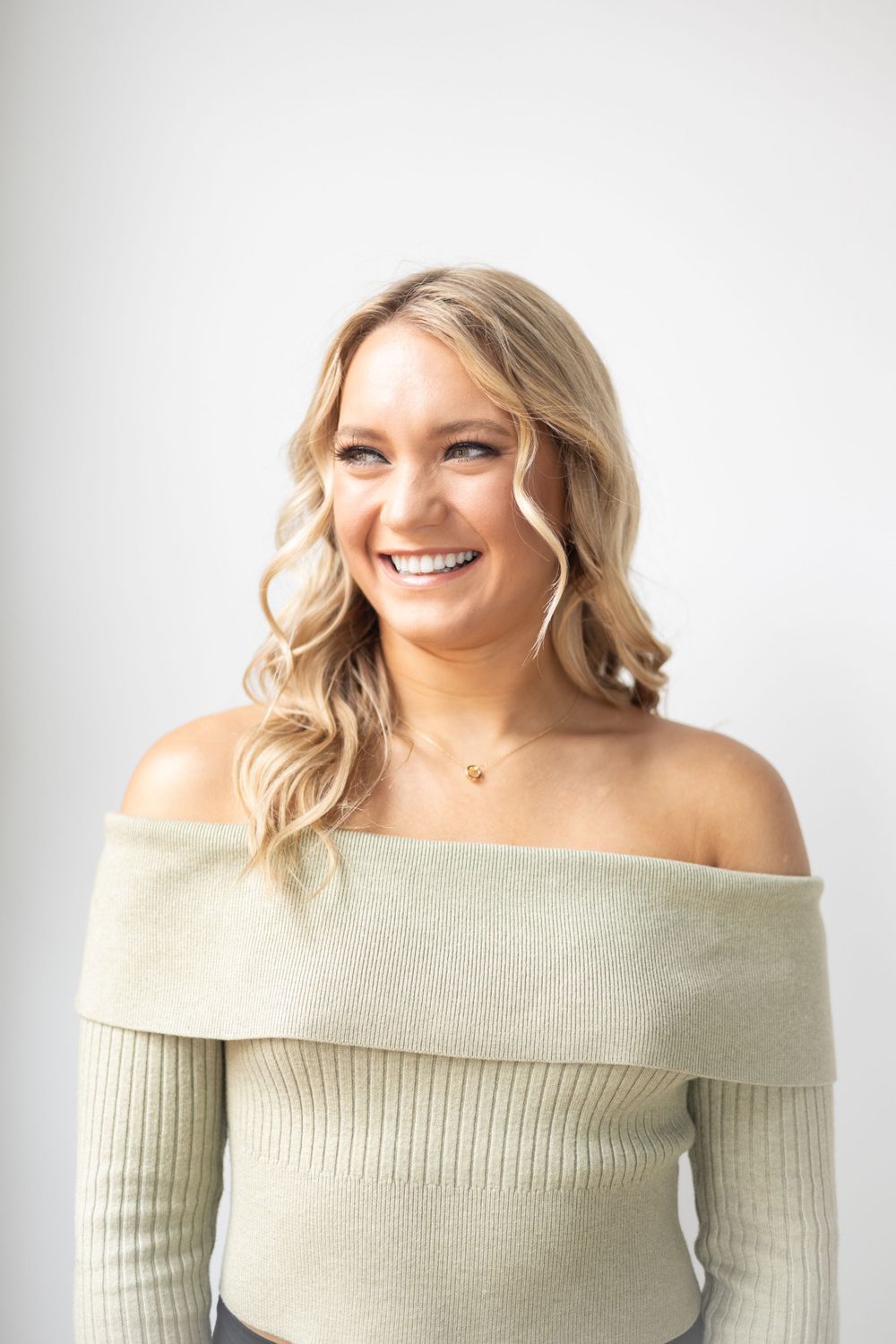 A person wearing an off-shoulder cream ribbed sweater smiles brightly against a white background.