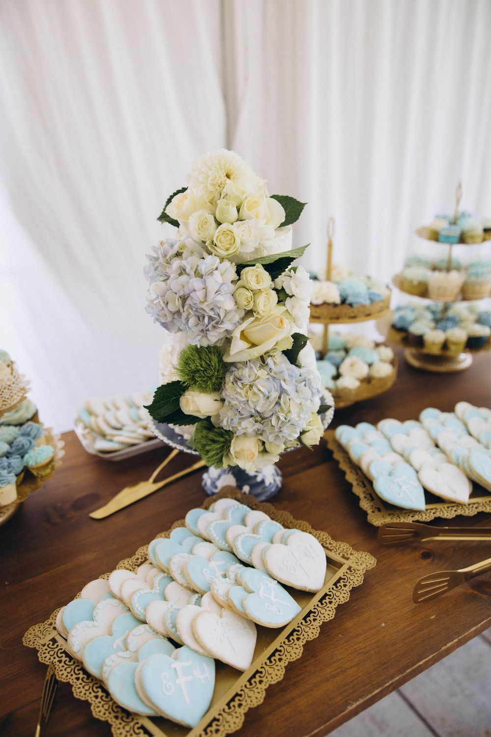 Elegant dessert table with white hydrangeas, cream roses, and light blue macarons arranged on gold doily platters.