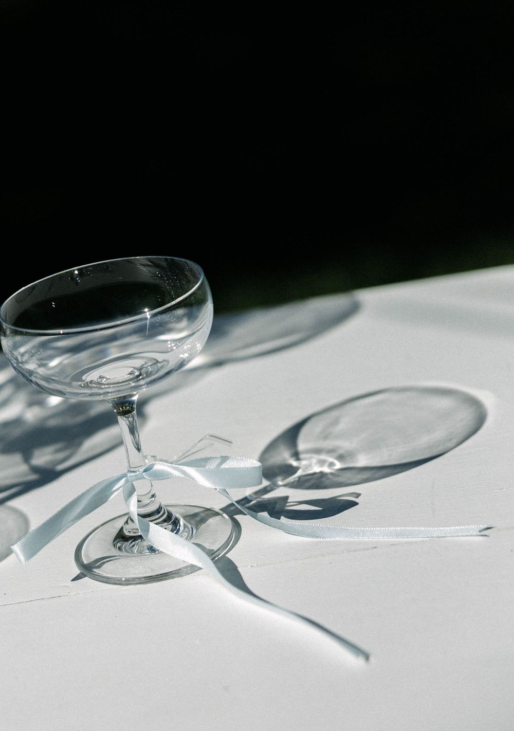 A clear coupe champagne glass casts dramatic shadows on a white surface in natural lighting.