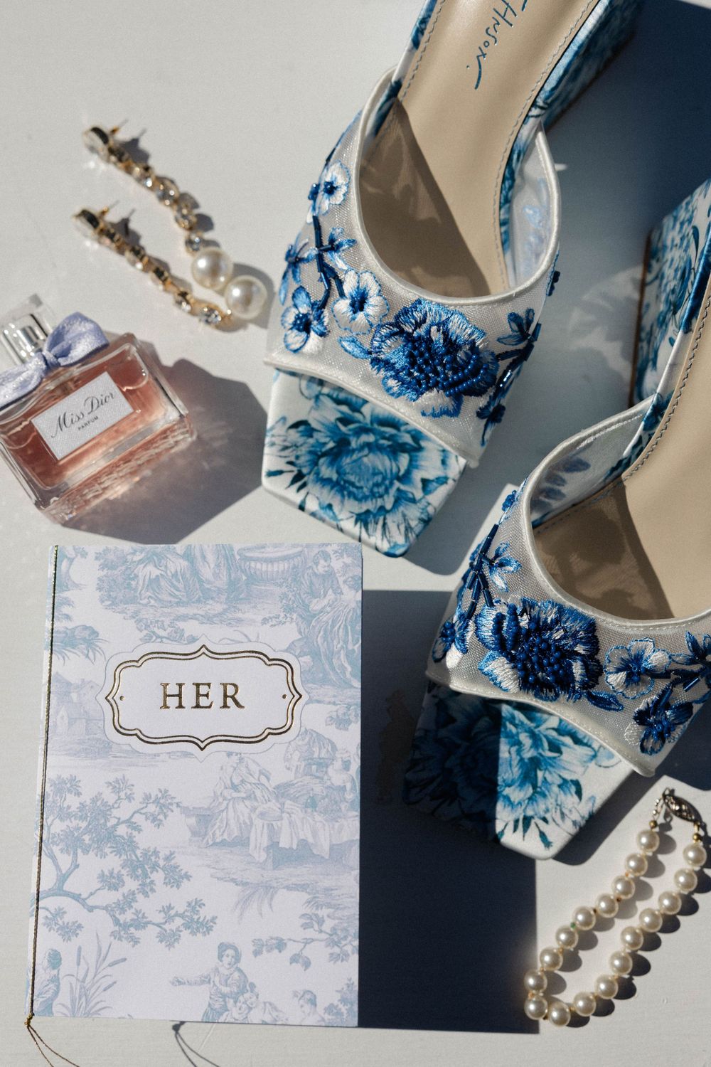 Floral blue and white wedding shoes arranged with perfume bottle, pearls and a 'HER' vow card on dark surface.