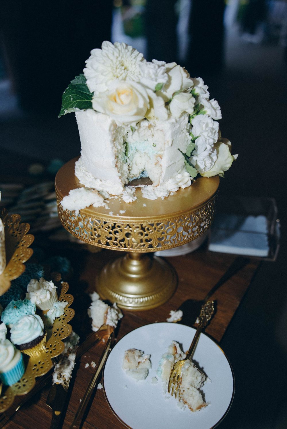 A white wedding cake with fresh roses on a gold ornate cake stand, with a slice removed and placed on small plate below.