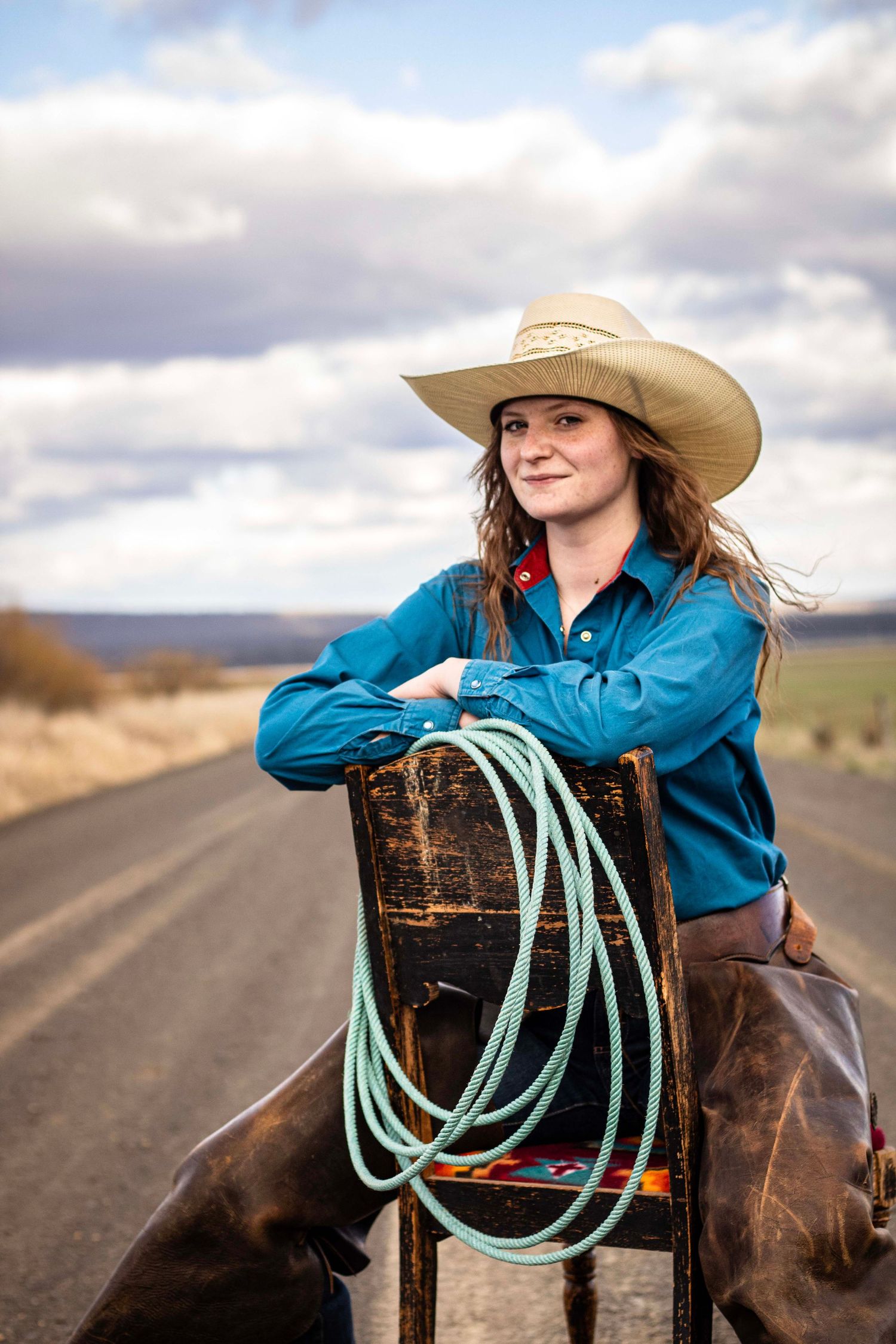 young woman sits on backwards chair with rope, chaps, and cowboy hat. behind her is a long dirt road, hills, and a cloudy sky