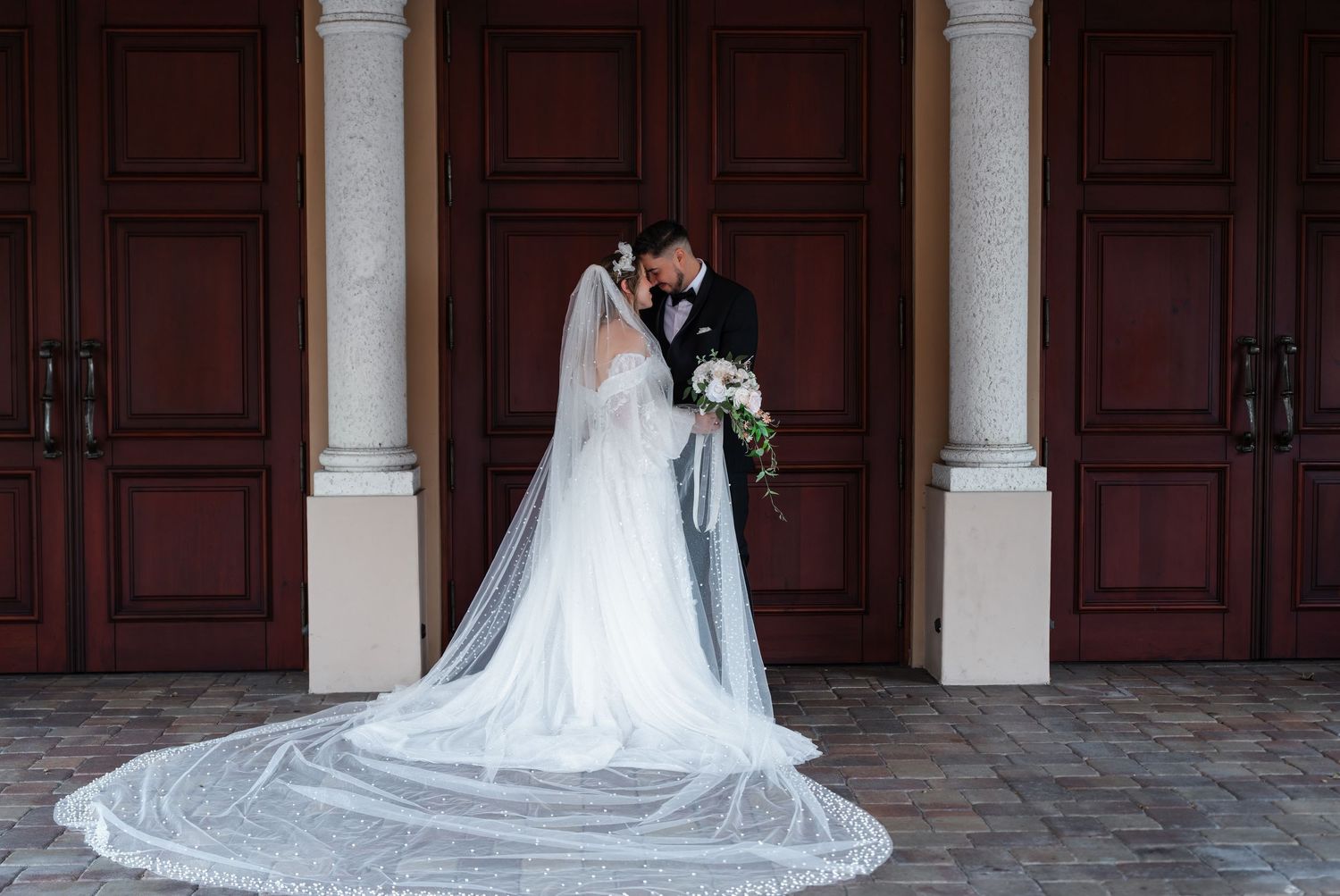 Elegant bride and groom share an intimate moment in front of grand wooden doors. The bride’s long, beaded veil cascades onto the cobblestone
