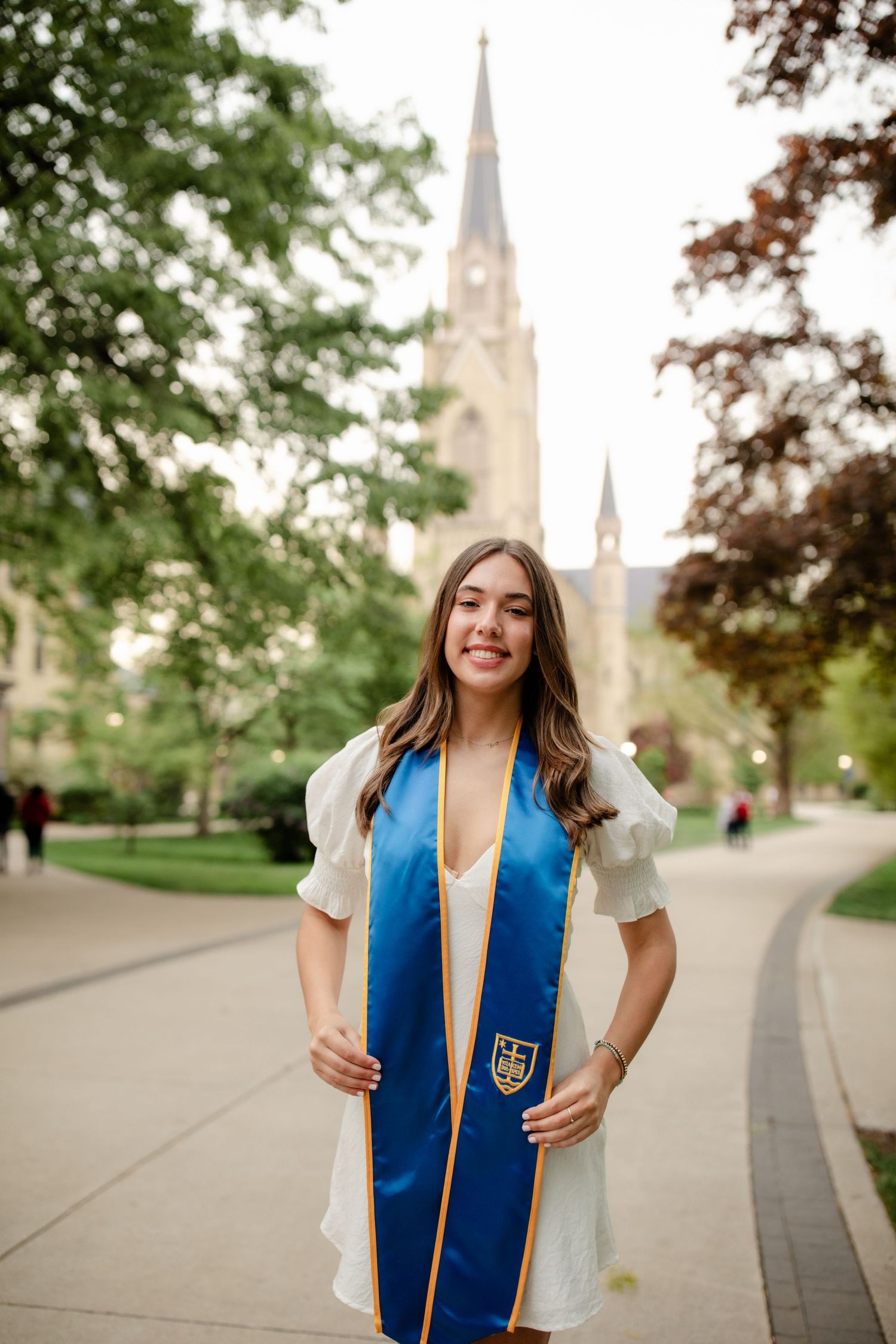 Graduate in white dress with blue stole standing on campus with cathedral spire in background.