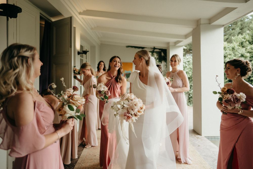 A group in blush pink bridesmaid dresses gather with the person in white on a covered porch.