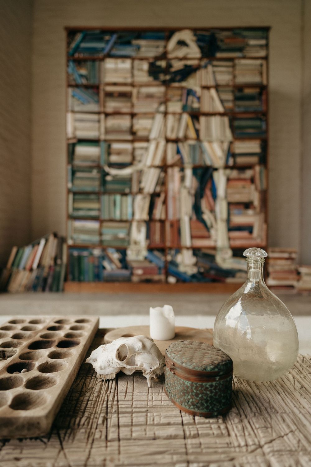 Vintage still life with glass bottle, wooden tray and decorative items against a wall of stacked books.