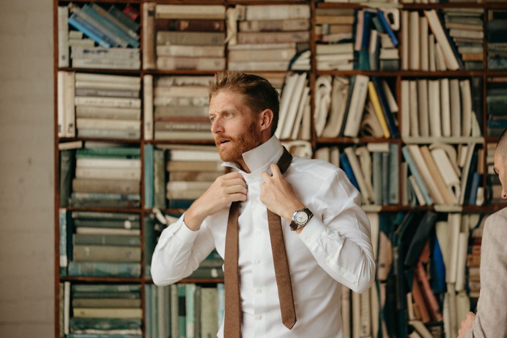 A person adjusts their brown tie while getting dressed in a room with bookshelves in the background.