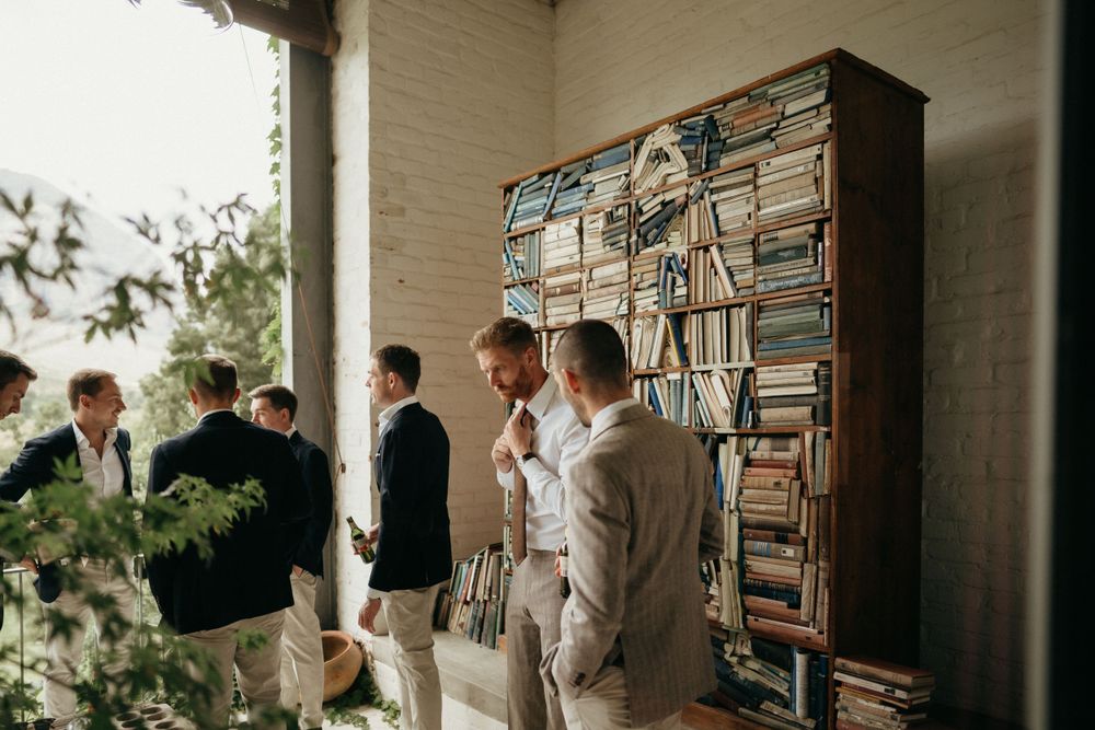 Groomsmen getting ready in casual setting with artistic bookshelf wallpaper backdrop.