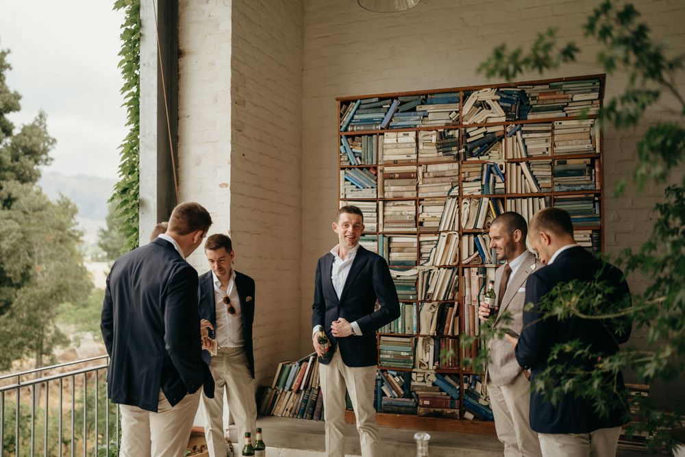 Wedding party members in navy blazers chatting on an elegant porch with climbing vines.