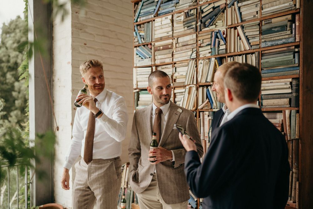 Groomsmen in light suits having drinks and conversation near wall of books.