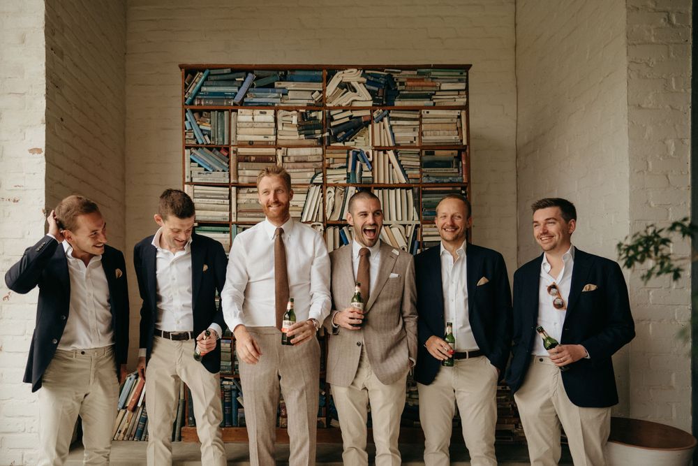 Groomsmen in mixed formal attire posing against textured wall with bookshelves.